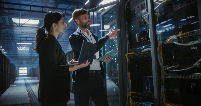 Two IT specialists use a laptop and tablet to inspect and analyze server racks in  data center, doing hardware diagnostics, network connectivity chekup, system equipment management