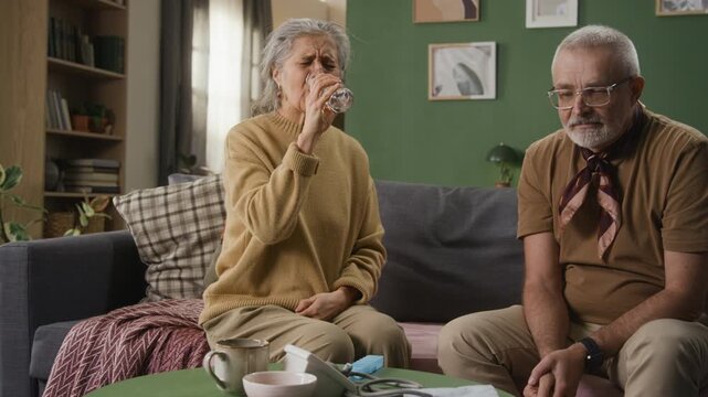 Caring husband giving glass of water to senior wife who taking pill screwing up her face while both sitting on couch