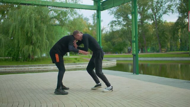 Two white men sparring under pavilion, park lake backdrop, casual sportswear, grappling drills, takedown attempt, clinch control, focused expressions, green trees, overcast light, training atmosphere