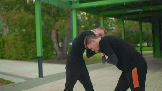 two white men performing hip throw under green pavilion, instructor guiding trainee through leverage and momentum technique, close contact, gripping and footwork coordination, park walkway and autumn