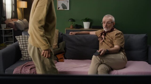 Elderly woman sitting down on couch next to husband and inviting white dog to join them, then taking book and reading while man using remote control