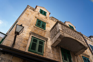 Old stone house facade with green shutters and a decorative balcony, showing elegant Mediterranean architecture against a clear blue sky.