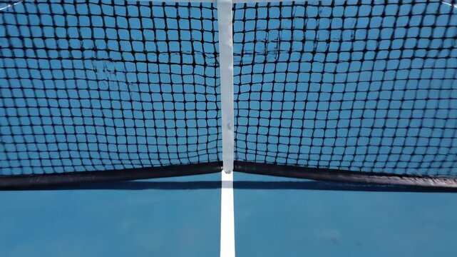 Aerial dolly shot moving forward over the net of an empty blue and green hard court in an outdoor sports facility, showing details of the playing surface and markings for a match