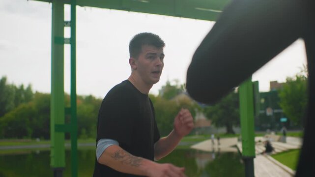 Male boxer dodging incoming kick outdoors, defensive stance shifts as boot crosses foreground, tattoo detail on arm, urban park background with lake and walkway, gritty closeups reveal intensity