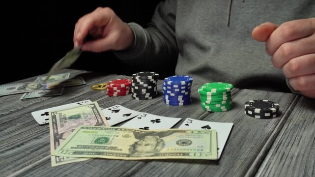 A gambler in a gray hoodie throws cash on playing cards on a wooden table and places a bitcoin coin. Poker chips are stacked around