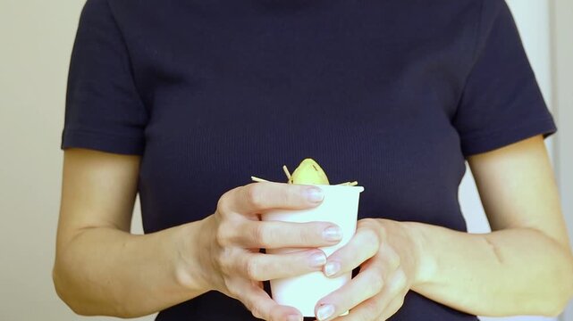Hands squeezing a paper cup with an avocado pit close to the camera before pulling it back. Represents hands-on home learning, step-by-step seed propagation, and interactive plant care practice.