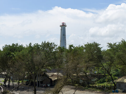 Aerial view of the stark white lighthouse rising above the lush green canopy under a bright sky, Mercusuar Samas, Bantul Regency, Indonesia.