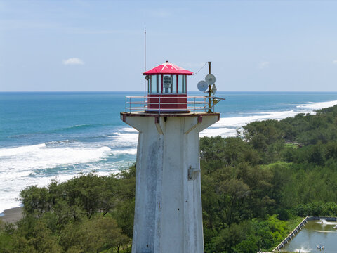 Aerial view of the Mercusuar Samas lighthouse standing tall against the backdrop of turquoise waves and lush greenery, Bantul Regency, Daerah Istimewa Yogyakarta, Indonesia.