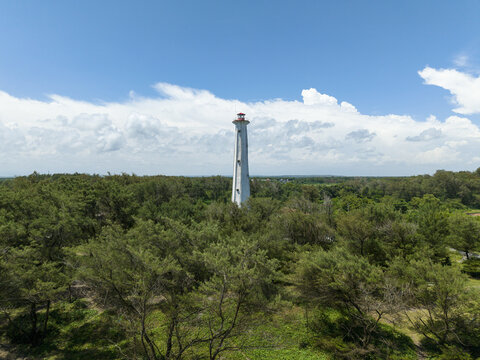Aerial view of a striking white lighthouse piercing through a sea of lush green trees under a vast, bright blue sky, Bantul Regency, Daerah Istimewa Yogyakarta, Indonesia.