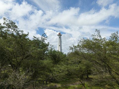 Aerial view of a towering white lighthouse piercing the sky, framed by lush green trees under a bright, partly cloudy sky, Bantul Regency, Daerah Istimewa Yogyakarta, Indonesia.