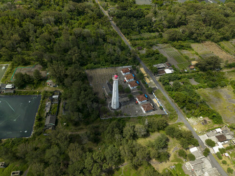Aerial view of a white lighthouse with a red top standing tall amidst the lush greenery and buildings near Mercusuar Samas, Bantul Regency, Daerah Istimewa Yogyakarta, Indonesia.