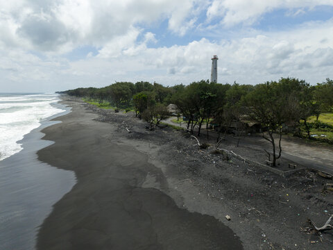 Aerial view of the lighthouse standing tall amidst the lush greenery along the dark sandy coastline, a stark contrast to the crashing waves, Bantul Regency, Daerah Istimewa Yogyakarta, Indonesia.