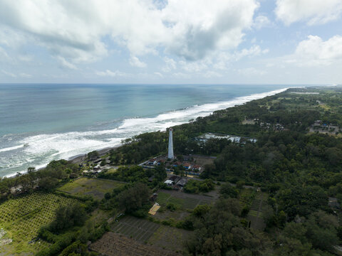 Aerial view of a towering lighthouse standing sentinel amidst lush greenery where the land meets the ocean's foamy embrace, Bantul Regency, Daerah Istimewa Yogyakarta, Indonesia.