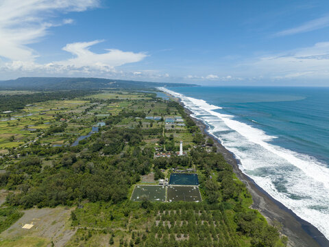 Aerial view of the coastline near Mercusuar Samas where the dark sand meets the frothy waves under a clear blue sky, Bantul Regency, Daerah Istimewa Yogyakarta, Indonesia.
