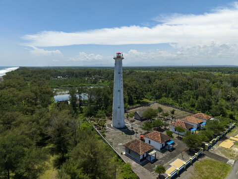 Aerial view of a striking white lighthouse piercing the skyline, contrasted against the lush green forest and the serene ocean, Mercusuar Samas, Bantul Regency, Indonesia.