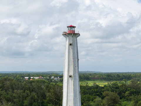 Aerial view of the stark white Mercusuar Samas lighthouse standing tall against a backdrop of lush green forest and cloudy skies, Bantul Regency, Daerah Istimewa Yogyakarta, Indonesia.