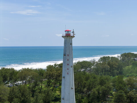 Aerial view of a majestic lighthouse named SAMAS, standing tall amidst verdant trees against the turquoise sea, under the vast blue sky, Bantul Regency, Daerah Istimewa Yogyakarta, Indonesia.