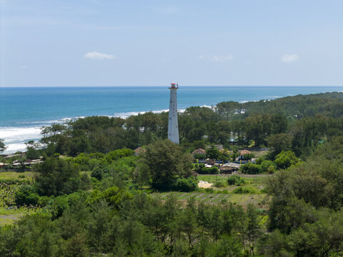 Aerial view of a white lighthouse standing tall amidst lush greenery, where the land meets the turquoise sea, creating a tranquil coastal scene, Bantul Regency, Daerah Istimewa Yogyakarta, Indonesia.