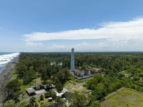 Aerial view of a towering white lighthouse standing proudly amidst lush green trees, its peak reaching towards the clear blue sky, Bantul Regency, Daerah Istimewa Yogyakarta, Indonesia.