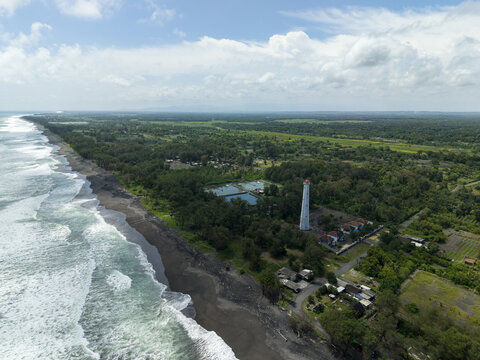 Aerial view of the stark white Mercusuar Samas lighthouse piercing through the lush green canopy along the dark sandy coastline, Bantul Regency, Daerah Istimewa Yogyakarta, Indonesia.