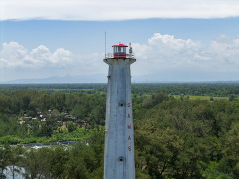 Aerial view of the tall, weathered Mercusuar Samas lighthouse rising above a sea of green trees under a vast blue sky, Bantul Regency, Daerah Istimewa Yogyakarta, Indonesia.