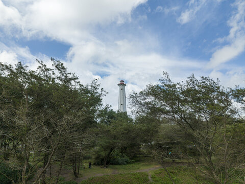 Aerial view of a tall, pristine lighthouse piercing through a canopy of lush green trees against a backdrop of soft, billowy clouds, Bantul, Daerah Istimewa Yogyakarta, Indonesia.