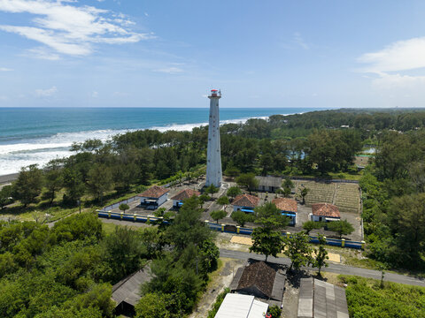 Aerial view of a striking white lighthouse rising amidst the lush greenery, contrasting with the dark sands and foamy waves of the sea, Bantul Regency, Indonesia.