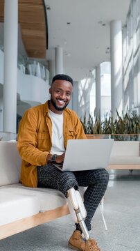 A confident professional with a prosthetic leg sits on a sofa using a laptop in a modern office space.