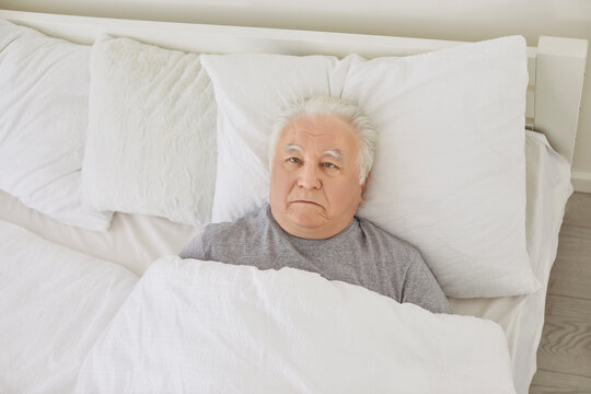 Senior man not sleeping. Unhappy old white haired man can't fall asleep lying awake in bed staring at ceiling with apathetic face expression late at night with light on. Insomnia sleeplessness concept