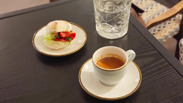 Espresso cup with appetizer on table, glass of water and tomato garnish, bright bistro setting, quick midday coffee after meeting