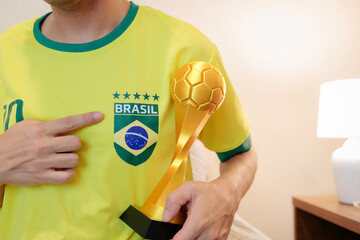Close-up de torcedor brasileiro apontando o escudo da camisa da seleção enquanto segura um troféu. Representa orgulho, vitória e paixão em uma competição internacional