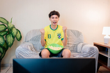 Torcedor brasileiro comemorando em casa enquanto assiste a um campeonato internacional de futebol pela TV. Ele veste a camisa da seleção brasileira, vibra no sofá e torce pela vitória do Brasil. © Marcio