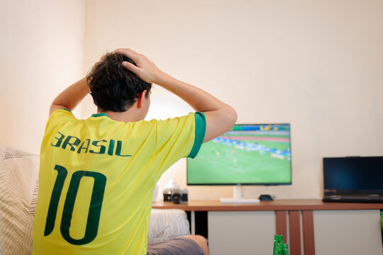 Torcedor brasileiro com a camisa da sele&ccedil;&atilde;o coloca as m&atilde;os na cabe&ccedil;a durante jogo tenso. De costas, assiste &agrave; TV em casa, expressando ansiedade, expectativa e esperan&ccedil;a de vit&oacute;ria do Brasil.