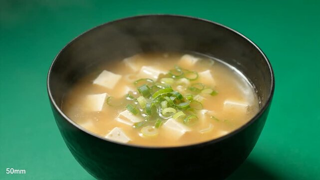 Steaming bowl of miso soup with tofu and green onions against a green backdrop