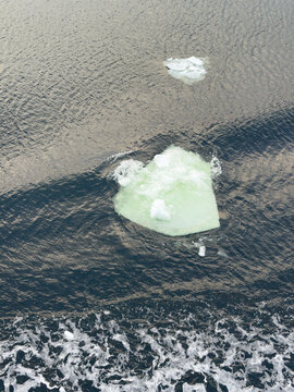 An iceberg drifts on the surface of dark water. The sunlight reflects off the water, showing ripples and soft waves around the ice. The setting is remote and cold