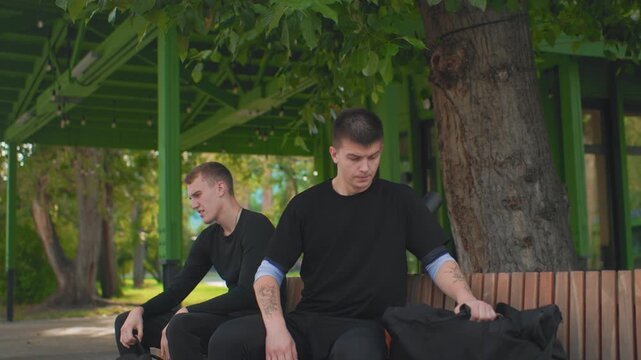 two caucasian men sitting on bench, tattooed forearm visible, urban park shelter, subdued expressions, distant gazes, subtle tension between friends, ambient green canopy, cinematic shallow focus,