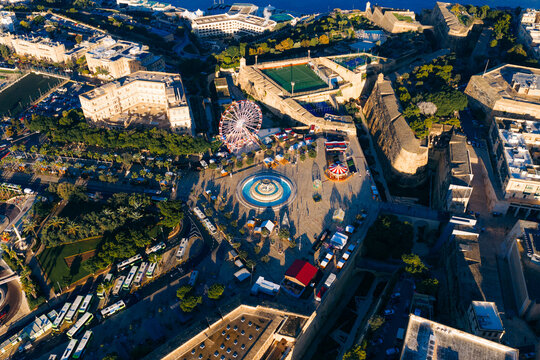 Aerial view of a vibrant city square featuring a Ferris wheel, fountain, and bustling market, framed by historic buildings and the azure sea, Valletta, Malta.