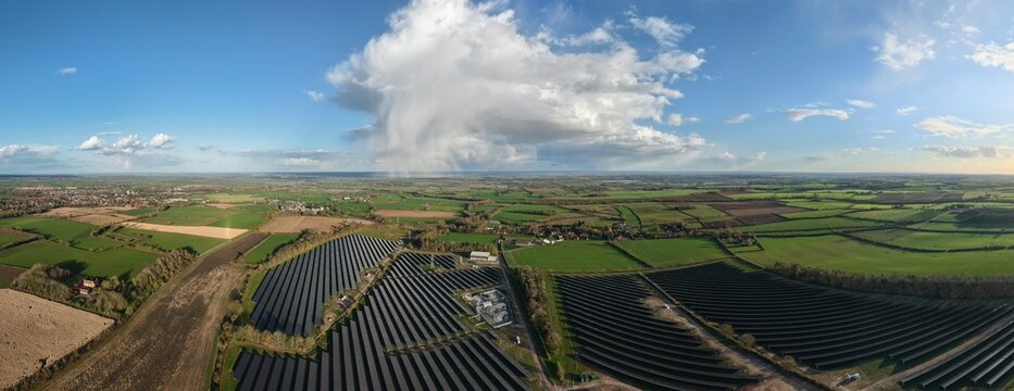 Aerial view of solar panels glinting under a vast sky punctuated by a dramatic cloud formation, contrasting with the patchwork of verdant fields, Halloughton, Southwell, United Kingdom.