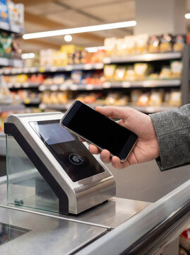 Shopper holds a smartphone over a payment terminal during self-checkout in a grocery store. Works well for mobile payment, retail technology, and automated checkout concepts.