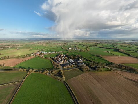 Aerial view of green fields and a cluster of buildings beneath a dramatic sky with a distant rain shower, Southwell, United Kingdom.