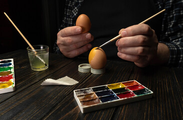 A person holds an egg and paints it with a brush. Watercolors are on the table along with a cup of water. The scene takes place in the evening with dim lighting © Віталій Б.