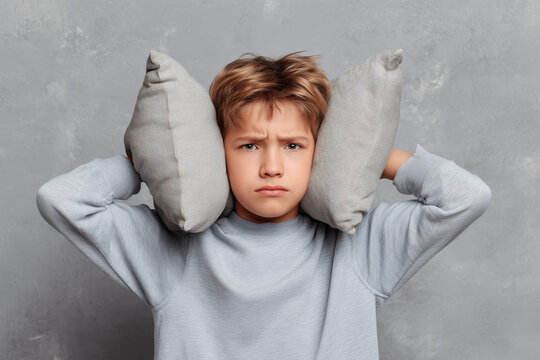 Boy covers ears with pillows while looking annoyed indoors