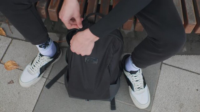 person on bench with helmet placing helmet into textured black backpack, hands working zipper, white sneakers resting on concrete sidewalk, fallen autumn leaves nearby, casual urban commuter scene