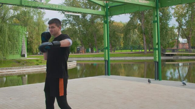 white male shadowboxing with gloves lakeside under green pavilion, focused footwork in orange shorts, calm pond backdrop, morning light, disciplined training routine, solo athlete preparing for match