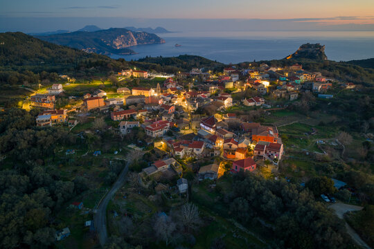 High angle drone view of Krini village streets and houses illuminated at dusk with Angelokastro castle overlooking the Ionian Sea in Corfu Greece