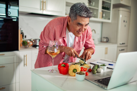 Smiling man cooking vegetables while following laptop recipe in home kitchen