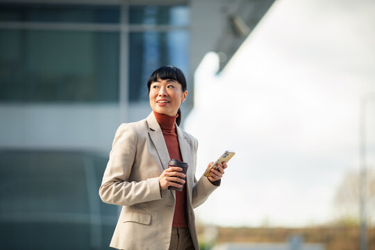 Smiling businesswoman with coffee and smartphone outside office building