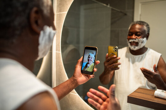 Senior man shaving during video call at home bathroom mirror
