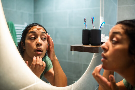 Young woman examining skin in bathroom mirror at home