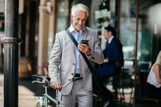 Senior businessman with bicycle using smartphone outside city cafe